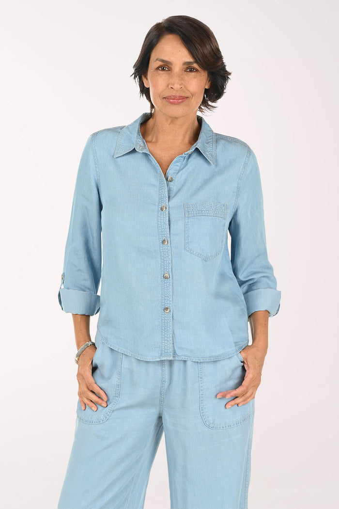 A smiling woman wearing a light blue denim button-up shirt and matching light blue denim pants, photographed against a plain light background.