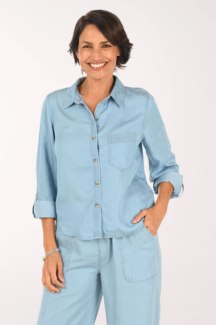 A smiling woman wearing a light blue denim button-up shirt and matching light blue denim pants, photographed against a plain light background.