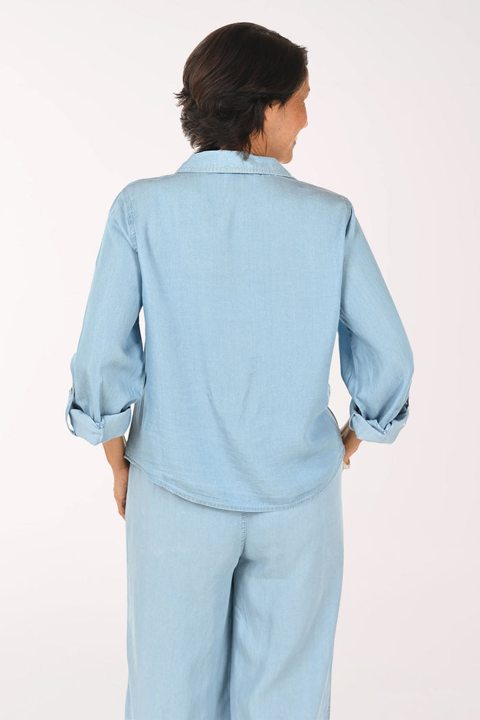 A smiling woman wearing a light blue denim button-up shirt and matching light blue denim pants, photographed against a plain light background.