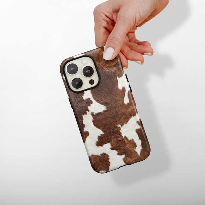 Cocoa Cowgirl Coconut Lane tough phone case with brown and cream cow print held in a hand against a white background.