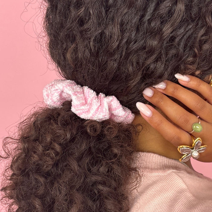 Curly-haired person wears a striped powder pink Coconut Lane towel scrunchie, rings, and pink nail polish on pink background.