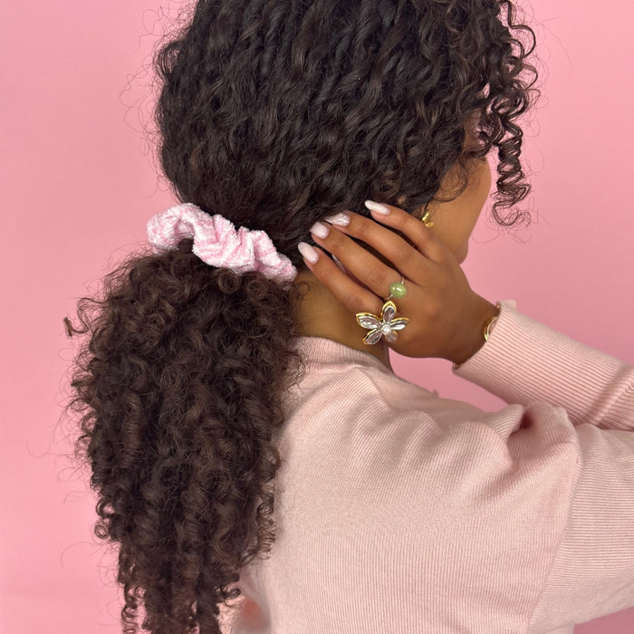 Woman with curly hair wears a striped powder-colored towel scrunchie by Coconut Lane, set against a pink background.