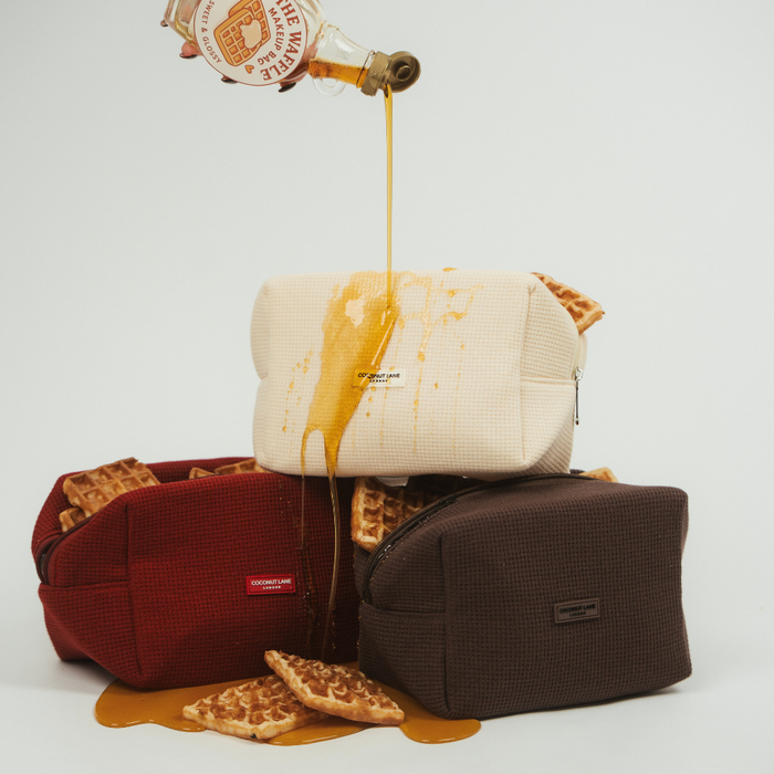 Three cream waffle-textured makeup bags stacked with golden waffles and syrup drizzled over them.