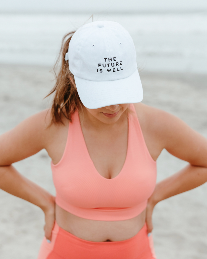 Woman wearing a white Wellness Month Dad Hat - Future is Well - White | Wellness Month with black text, standing on a beach with hands on hips, dressed in coral sportswear.