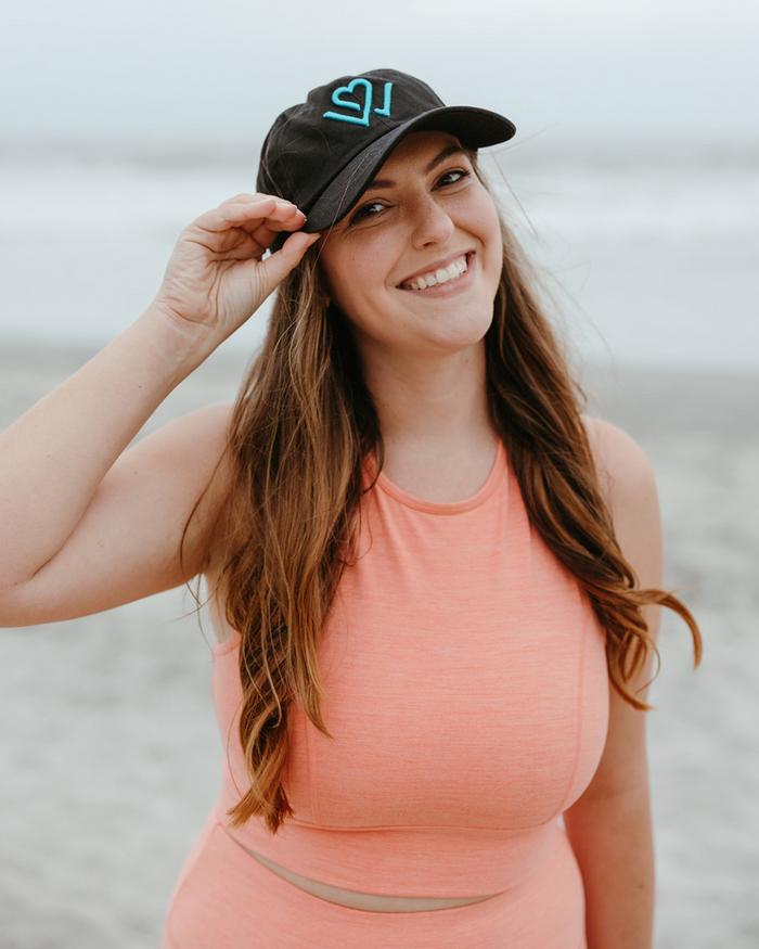 Smiling woman wearing a Wellness Month Logo Dad Hat - Black w/Blue Logo | Wellness Month at the beach, dressed in coral activewear, showcasing casual wellness style.