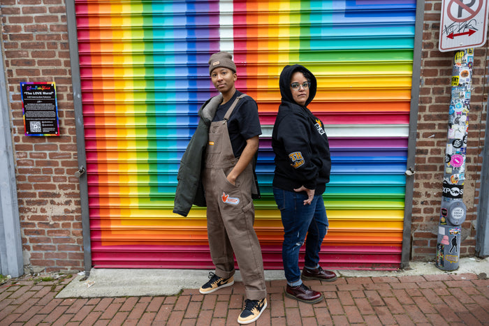 Two individuals stand back-to-back in front of a vibrant rainbow mural painted on a metal garage door. The person on the left wears brown overalls with a "Collective Action" patch, a black t-shirt, a brown beanie, and tan-and-black sneakers, with a green jacket draped over one shoulder. The person on the right wears a black hoodie with the "WE/US together" design on the front, blue jeans, and burgundy boots, with the hood up. They both have confident expressions, representing unity, empowerment,