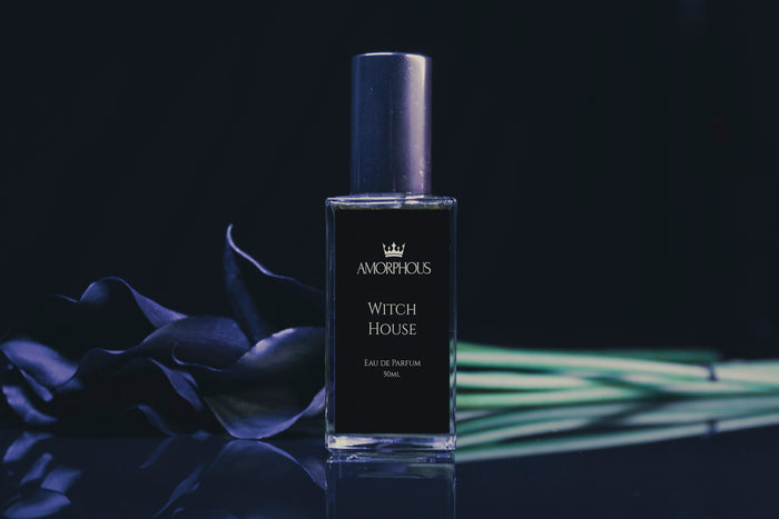 A bottle of Amorphous Witch House Eau de Parfum stands on a reflective dark surface against a black background, styled with dark blue calla lilies and green leaves.